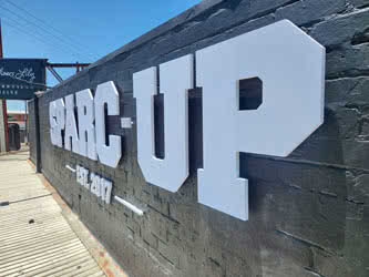 Closeup front/right view of large white lettering 'SPARC - UP', a business sign, fixed onto a black-painted brick wall, sunny clear day