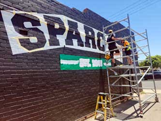 Worker on a scaffold painting business signage onto a black-painted brick wall, outside on a sunny day