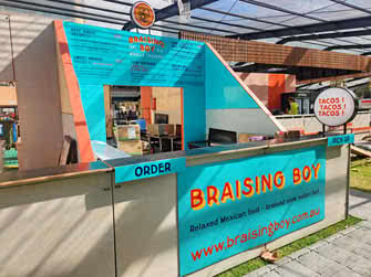 Food stall showing menus and business name sign, belonging to Braising Boy Tacos, front view from left