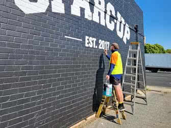 Worker next to and standing on another ladder, painting finishing touches of business signage onto a black-painted brick wall, outside on a sunny day